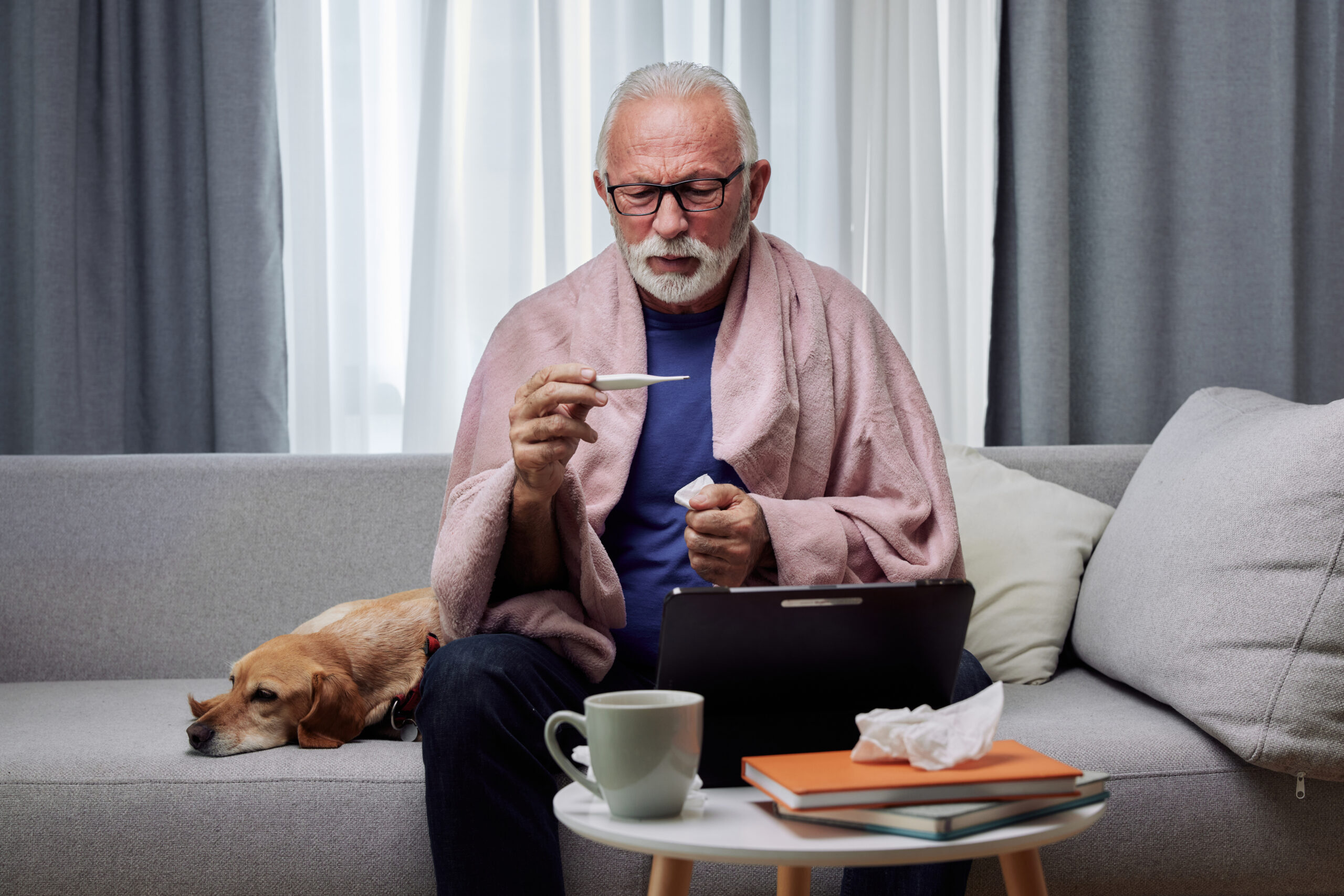 Sick elderly man checking his temperature suffering from seasonal flu or cold and using digital tablet for entertainment or online doctor consultation. Ill senior feel unhealthy with influenza at home