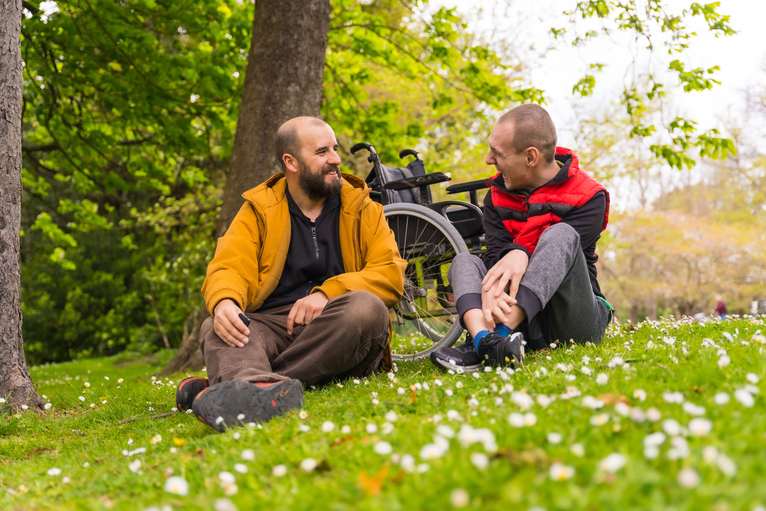 A young man with a disability and a friend sitting on the grass of a public park in the city, talking and laughing. A wheelchair sits in the background.