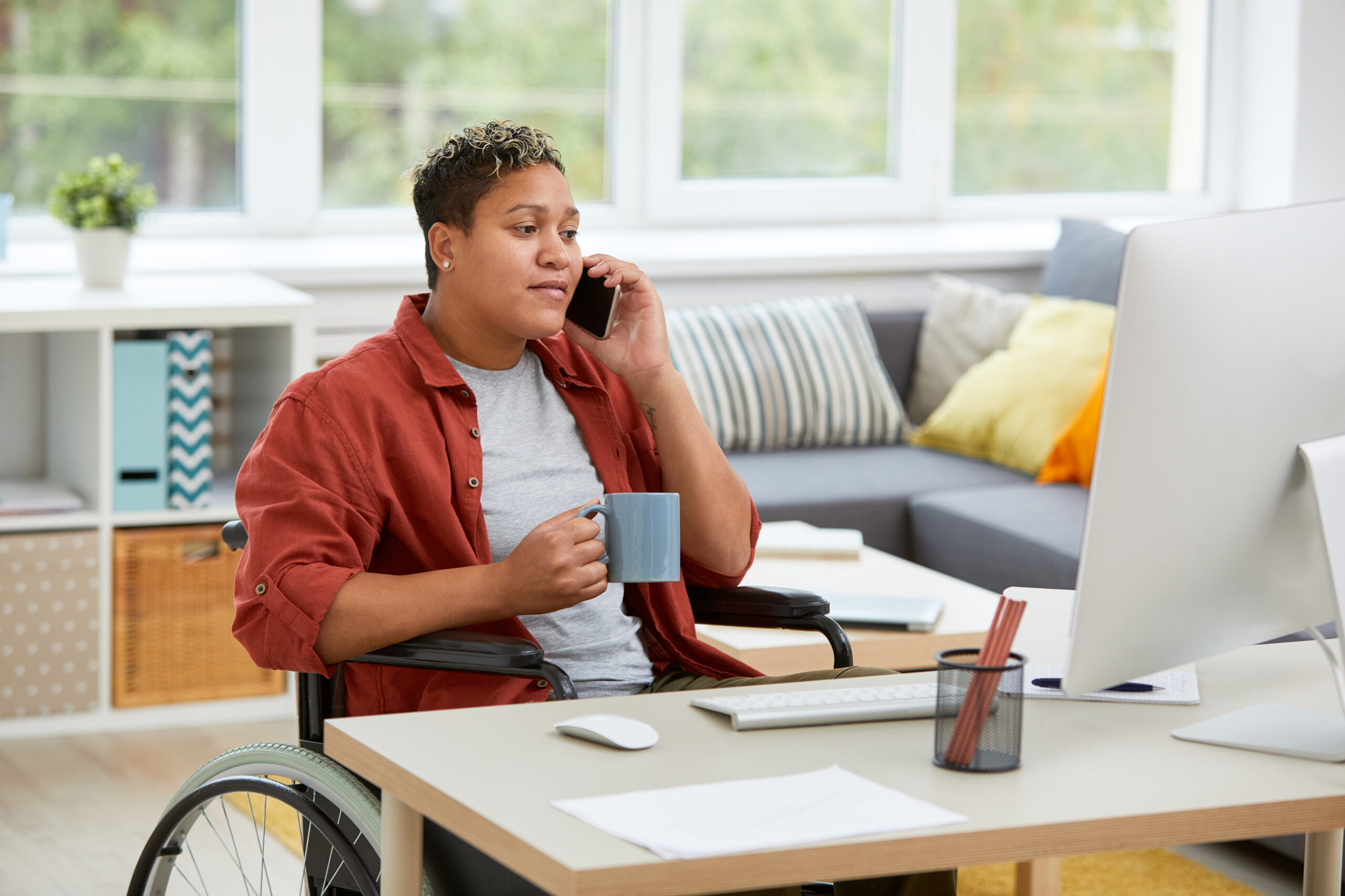 Woman sitting in her wheelchair at the table and looking at computer monitor while talking on mobile phone sharing why Medicaid Funding Access is important to her Self-Direction journey.